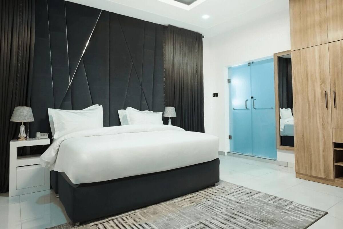 A modern bedroom with a large bed, dark headboard, bedside lamps, a wardrobe, and a blue door.