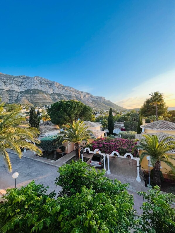 A residential area with a mountain backdrop, featuring a paved driveway, a white fence, and lush greenery.