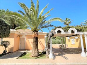 Un edificio con una palma in primo piano, una zona porticata e un ingresso con cancello.