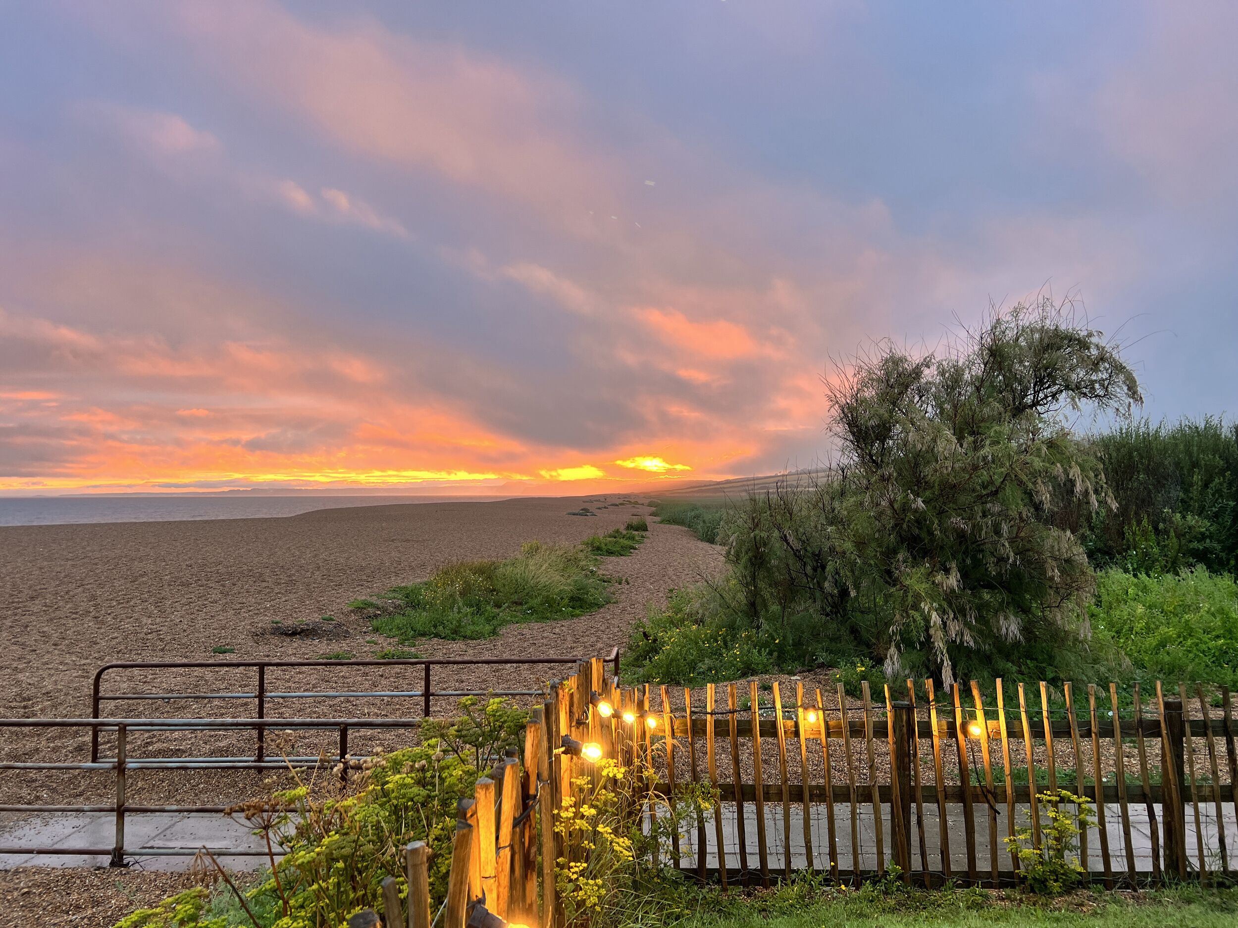 A sunset view over a beach with a wooden fence and a metal railing in the foreground.