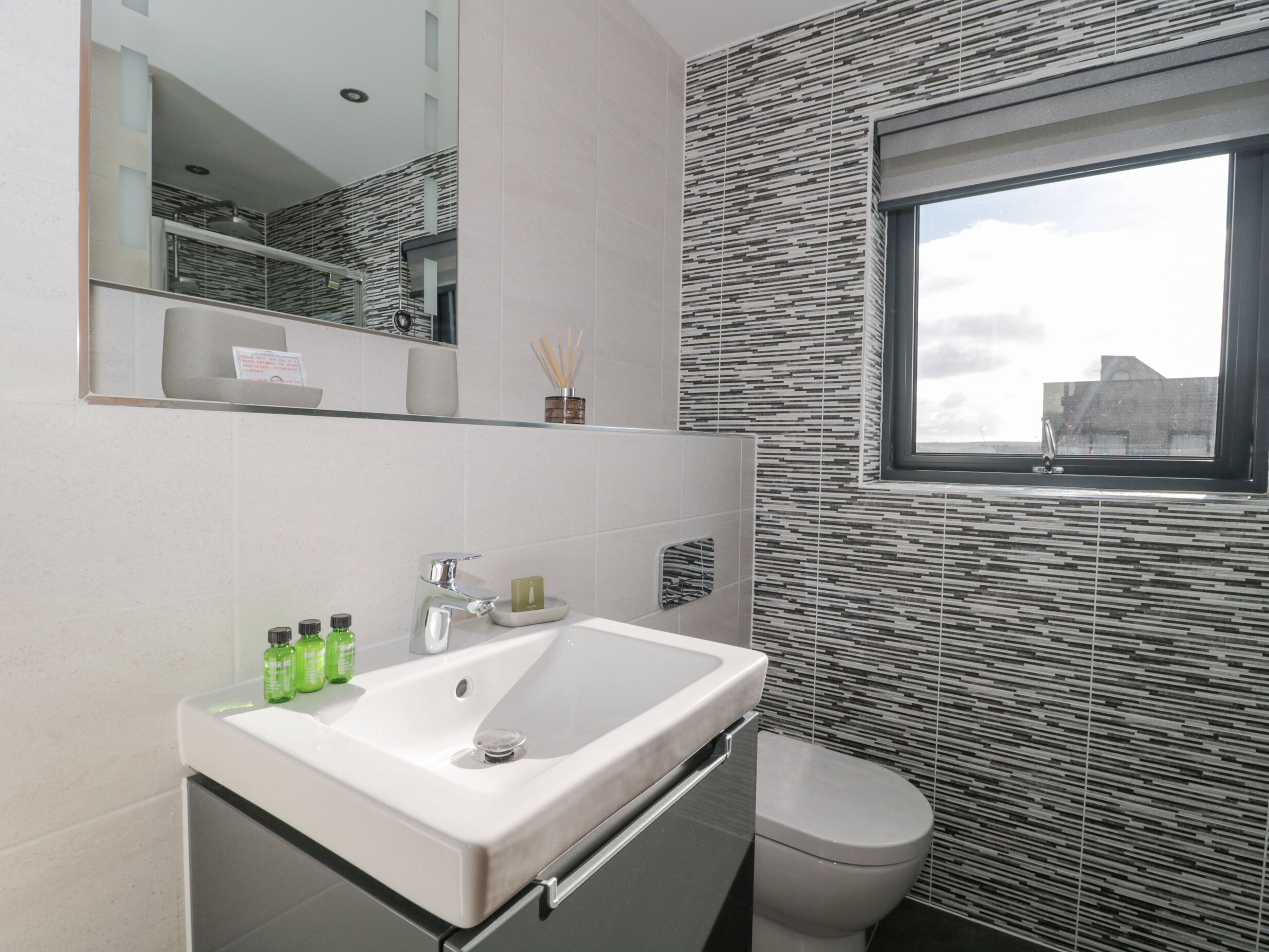Un bany modern amb un lavabo blanc, un mirall a sobre i un vàter. Hi ha una finestra amb vista a edificis.