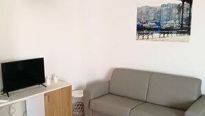A living room with a flat-screen TV, a white sofa, a wooden cabinet, and a painting of a cityscape on the wall. - settimo sole affittacamere (Taranto)