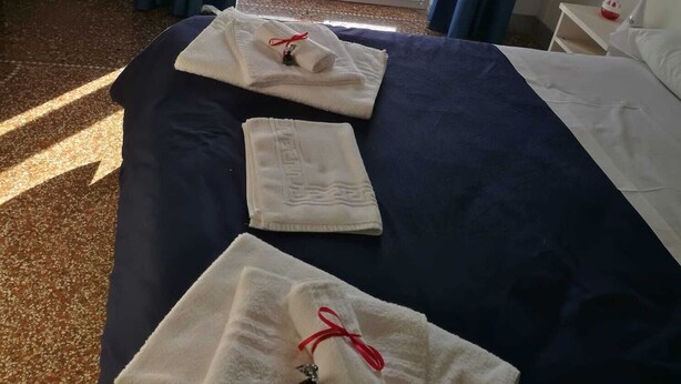 A hotel room with a bed, two towels, a nightstand, a lamp, and a view of buildings through a glass door. - settimo sole affittacamere (Taranto)