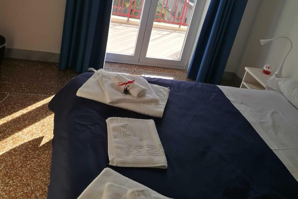 A hotel room with a bed, two towels, a nightstand, a lamp, and a view of buildings through a glass door.