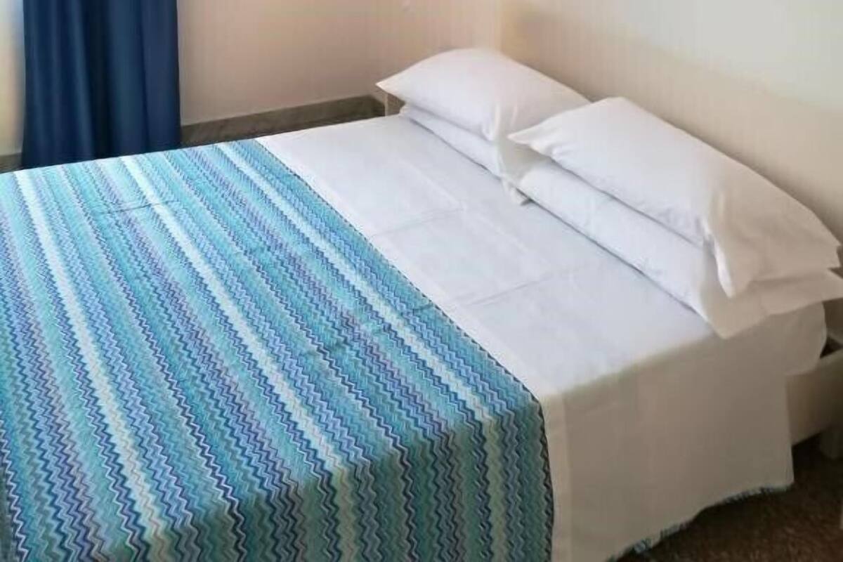A hotel room with a bed, a painting of a coastal scene, and a blue curtain.