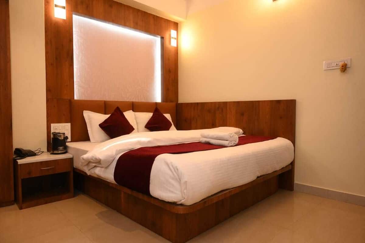A wooden bed with white and red bedding, a nightstand with a phone and a coffee maker, and a wall-mounted light fixture.