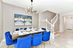 Un coin repas avec une table dressée pour six personnes, des chaises bleues, un lustre et deux tableaux abstraits accrochés au mur.