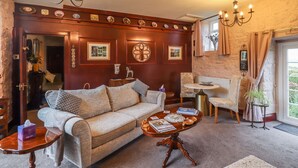 A cozy living room with a sofa, coffee table, and decorative items. There is a fireplace, a chandelier, and a window with curtains. - The Barn (Morpeth)