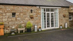 A stone building with a large window and a door, surrounded by potted plants and a stone bench. - The Barn (Morpeth)
