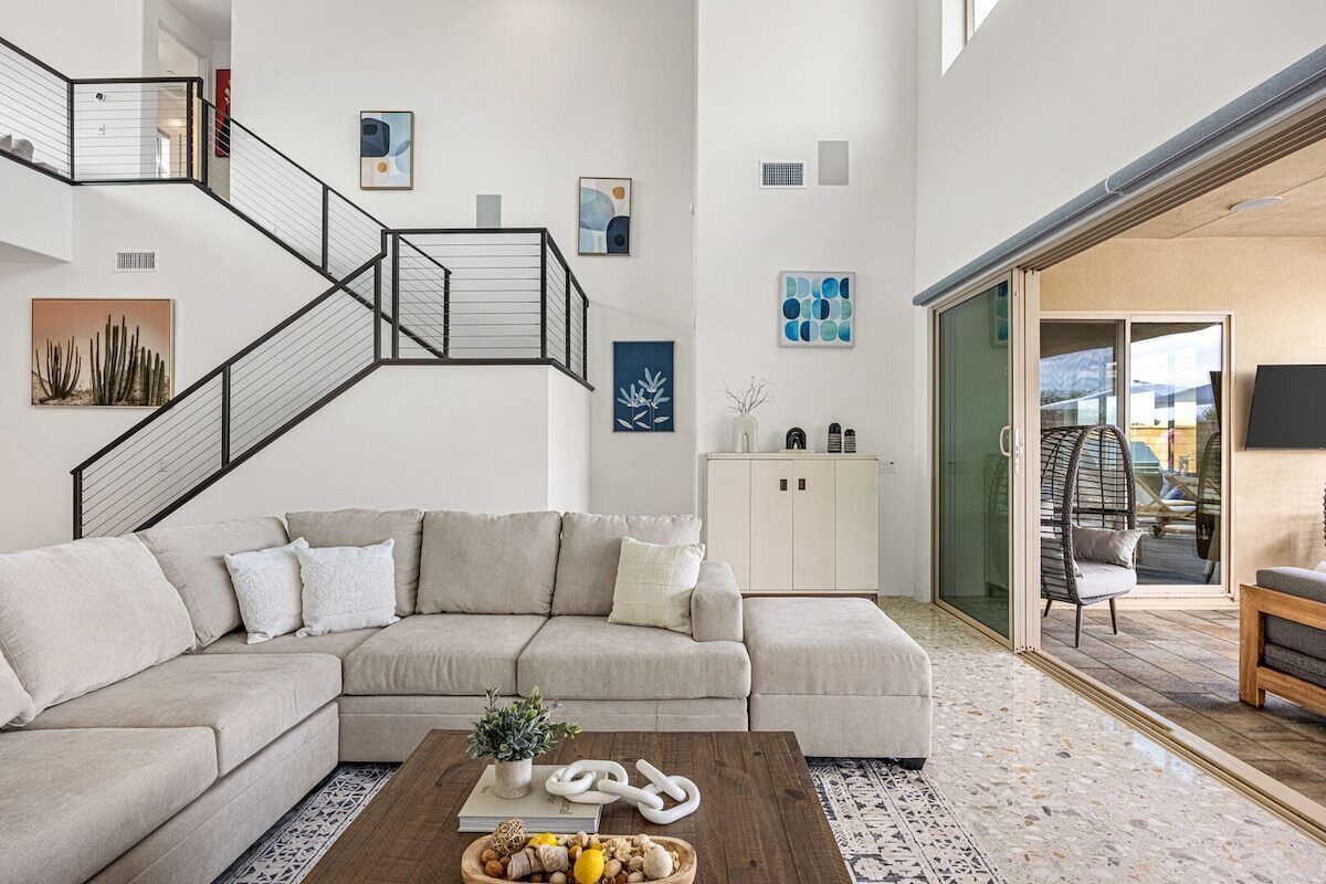 A modern living room with a sectional sofa, a coffee table with decorative items, a staircase with a railing, and a glass door leading to an outdoor area.