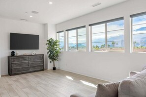 A spacious living room with a large flat-screen TV mounted on the wall, a dark wooden cabinet, a potted plant, and large windows offering a view of the outdoors. - Desert Oasis Retreat | EV Charger, GolfCourseViews (La Quinta)