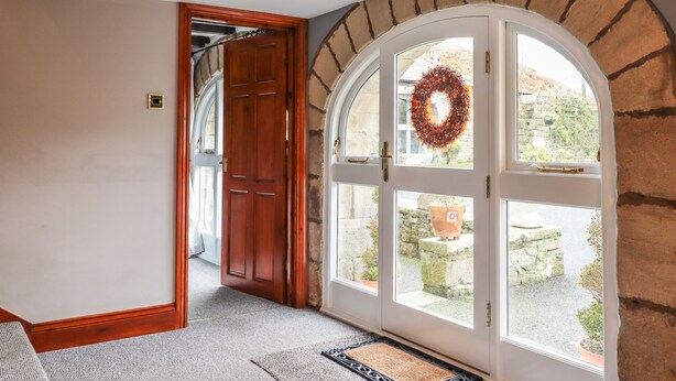 A hallway with a wooden door leading to a room, a large arched window with a wreath, and a stone wall. - High Shaftoe (Morpeth)