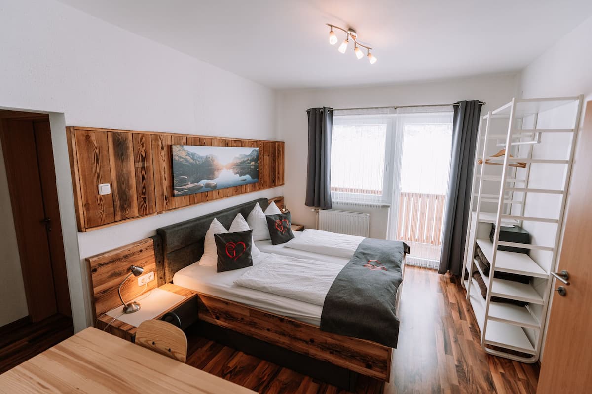 Une chambre à coucher avec une tête de lit en bois, un lit recouvert de linge de maison blanc, une table de chevet avec une lampe, une fenêtre avec des rideaux et une armoire intégrée.