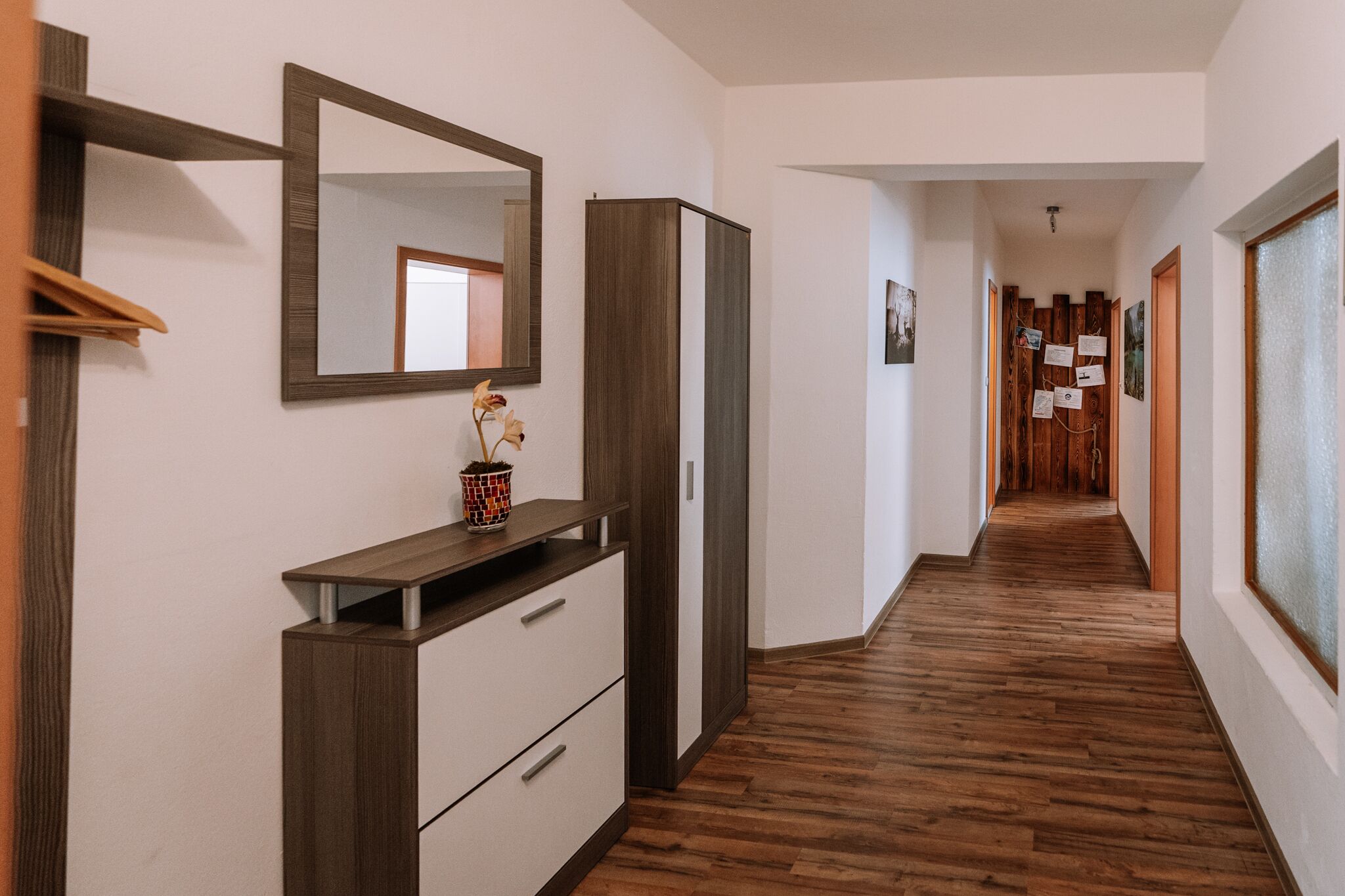 A hallway with wooden flooring, a mirror, a cabinet, and a potted plant.