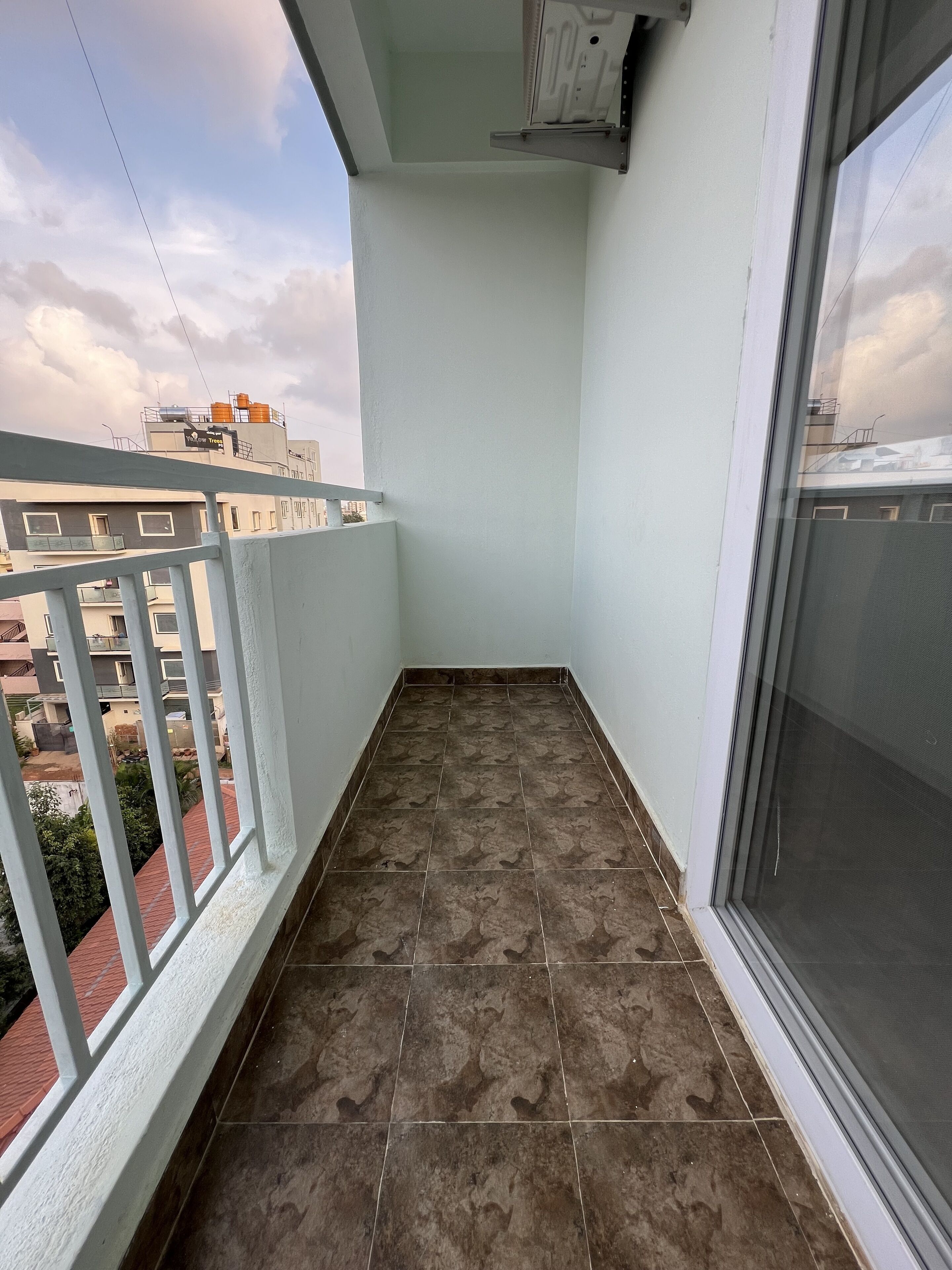 Un balcón con piso de baldosas, barandilla blanca y ventana con mosquitero.