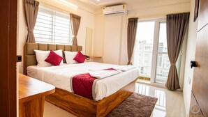 A modern bedroom with a large bed, a wooden bedside table, a view of the city through the window, and an air conditioning unit on the wall. - The Auberge - Boutique Hotel (Bengaluru)
