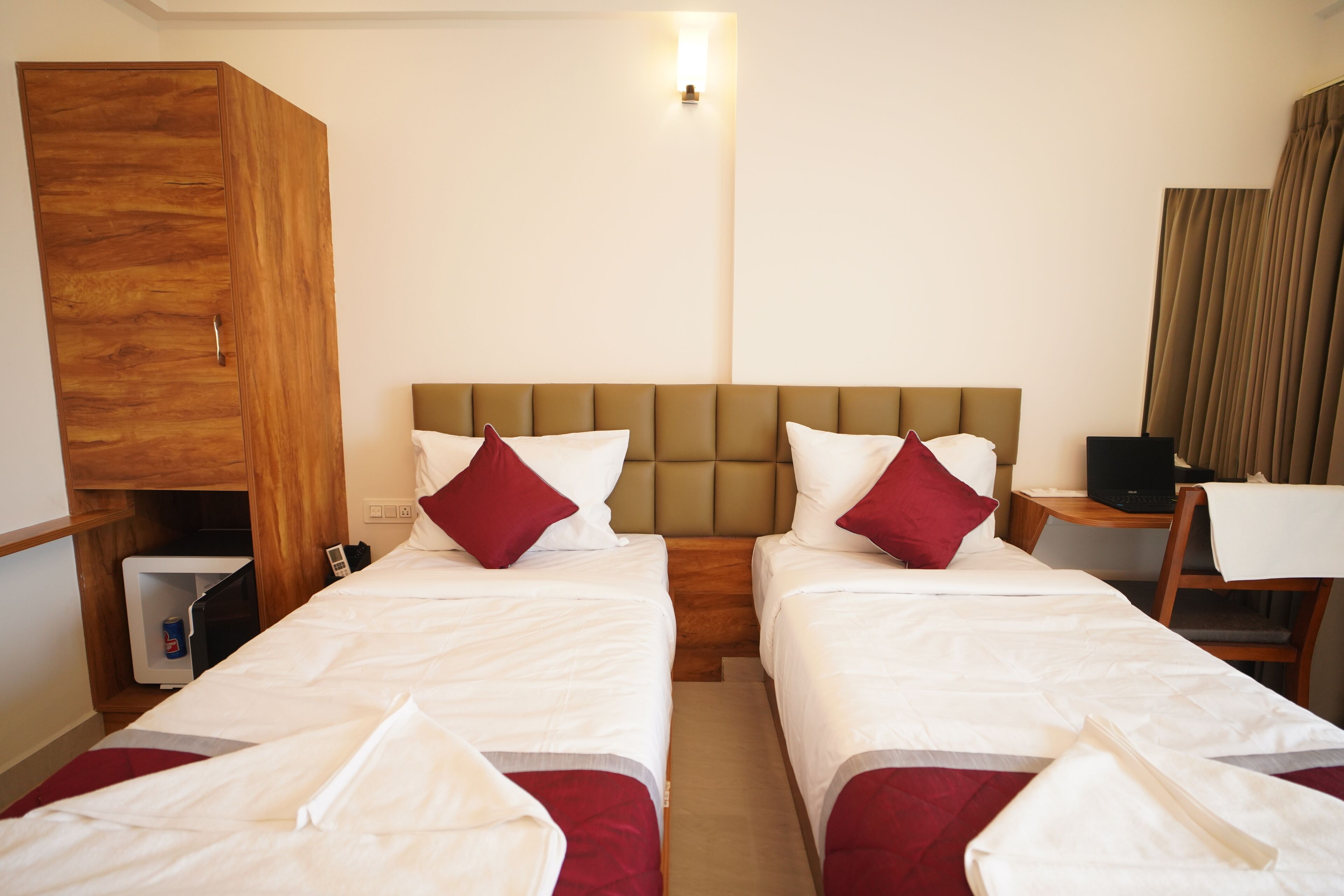 A hotel room with two single beds, a wooden wardrobe, a desk with a chair, and a TV.
