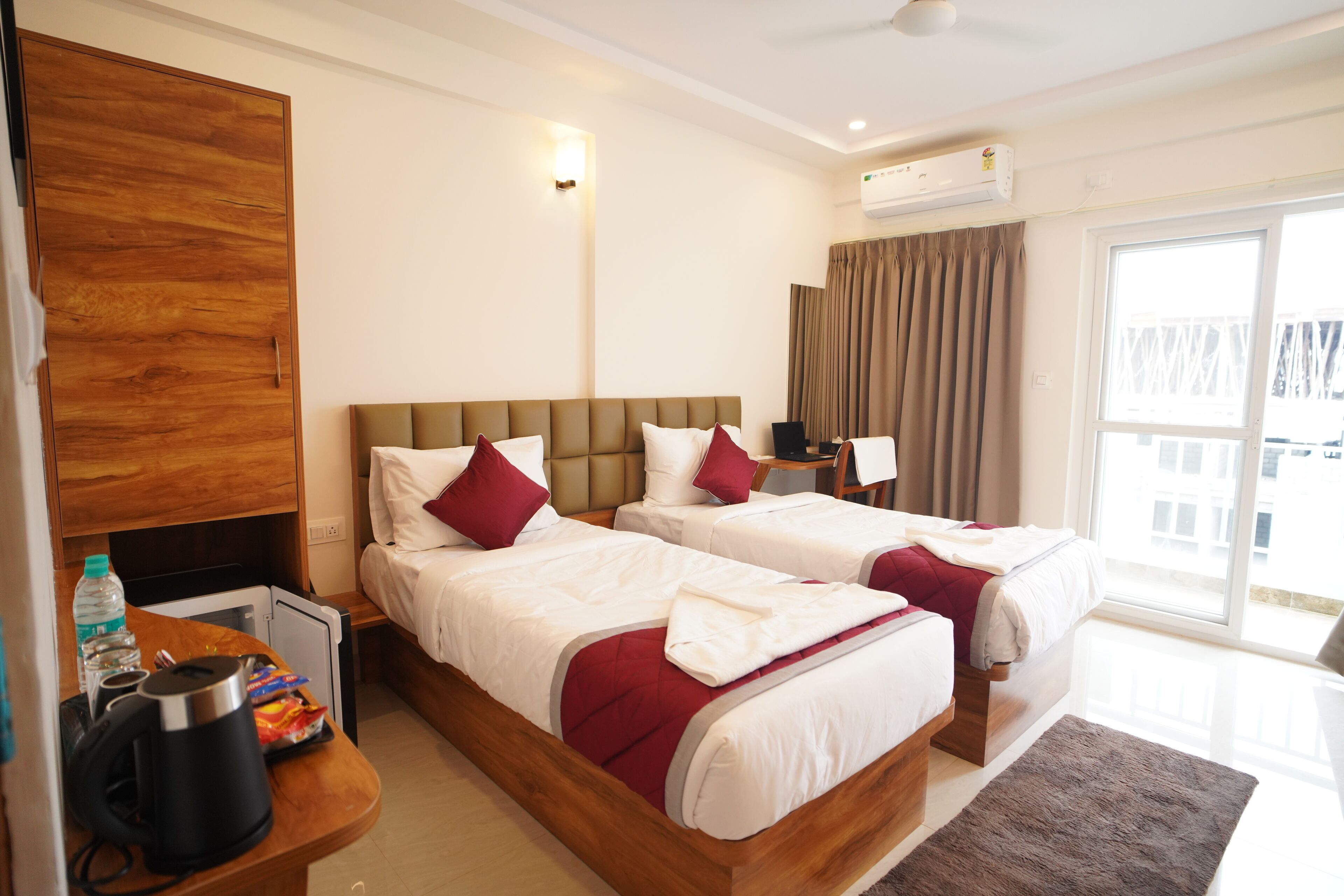 A hotel room with two beds, a wooden wardrobe, a desk with a kettle, and a large window with curtains.