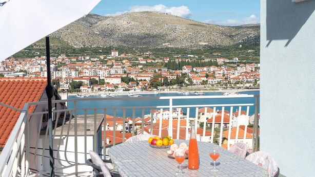A balcony with a table set for two, overlooking a coastal town with red-roofed buildings and a mountain in the background. - Anto - 3 bedroom apartment in Trogir (Trogir)