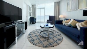 A modern living room with a flat-screen TV, a blue sofa, a round coffee table, and a large window offering a view. -  Cozy 1b With Balcony in Business Bay (Dubai)