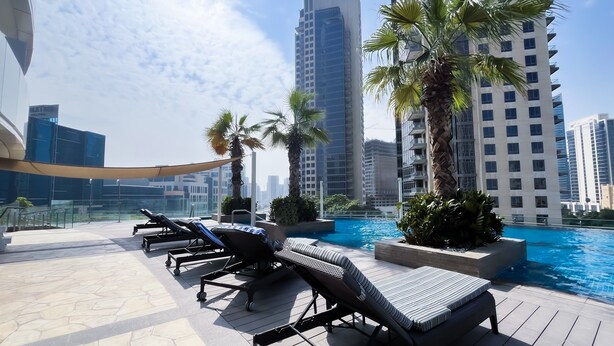 A rooftop pool area with lounge chairs and palm trees, surrounded by high-rise buildings. -  Cozy 1b With Balcony in Business Bay (Dubai)
