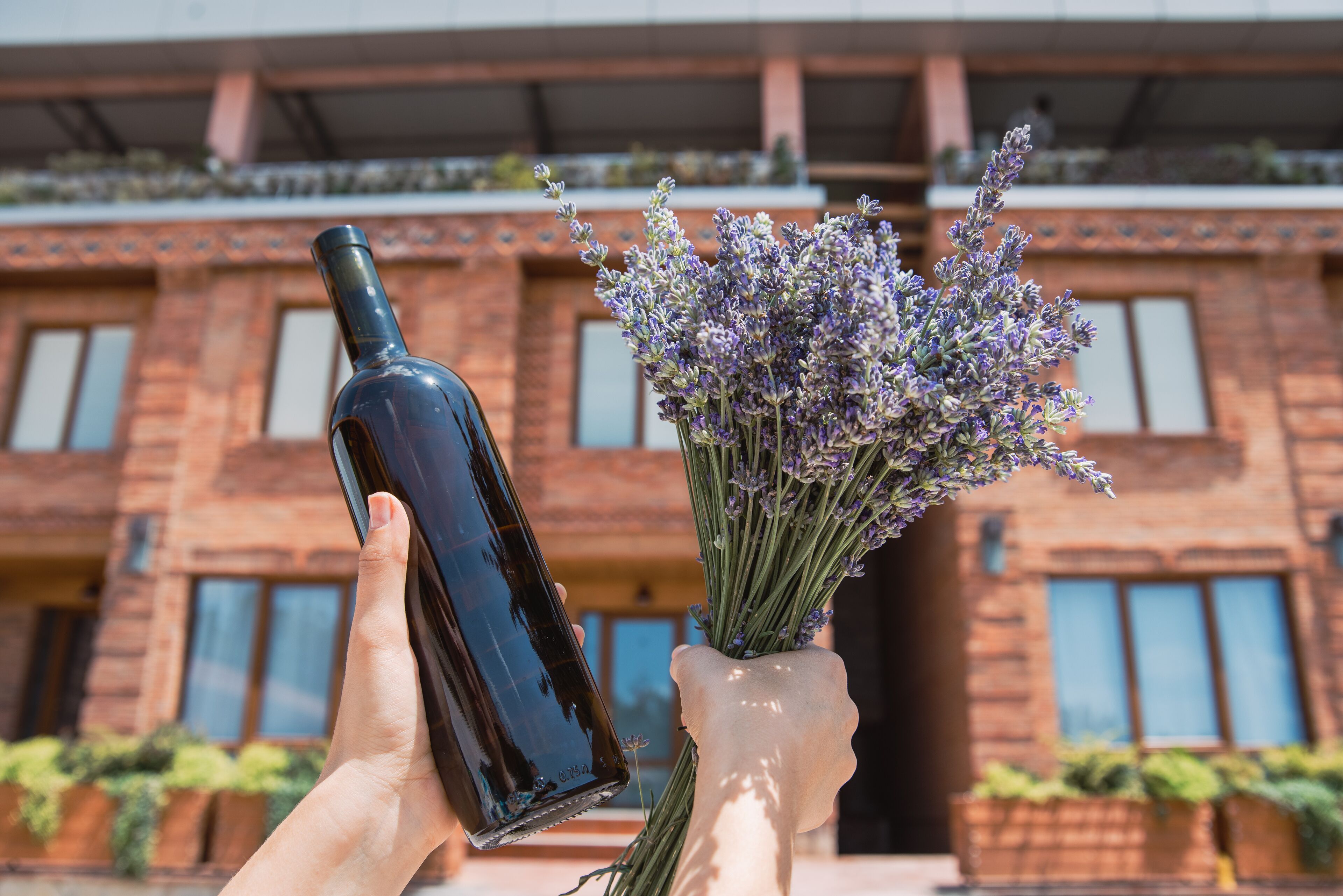 A hand holding a dark bottle and a vase with lavender flowers in front of a brick building with large windows.