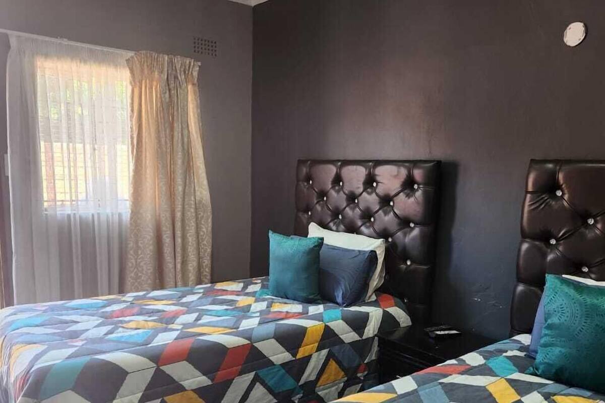 A hotel room with two beds, each with a geometric-patterned bedspread and matching pillows. The room has a dark-colored headboard and a window with curtains.