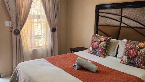 A neatly made bed with a red blanket, floral pillows, and a headboard with a geometric pattern. - Suits bar restaurant and guesthouse (Merafong City)