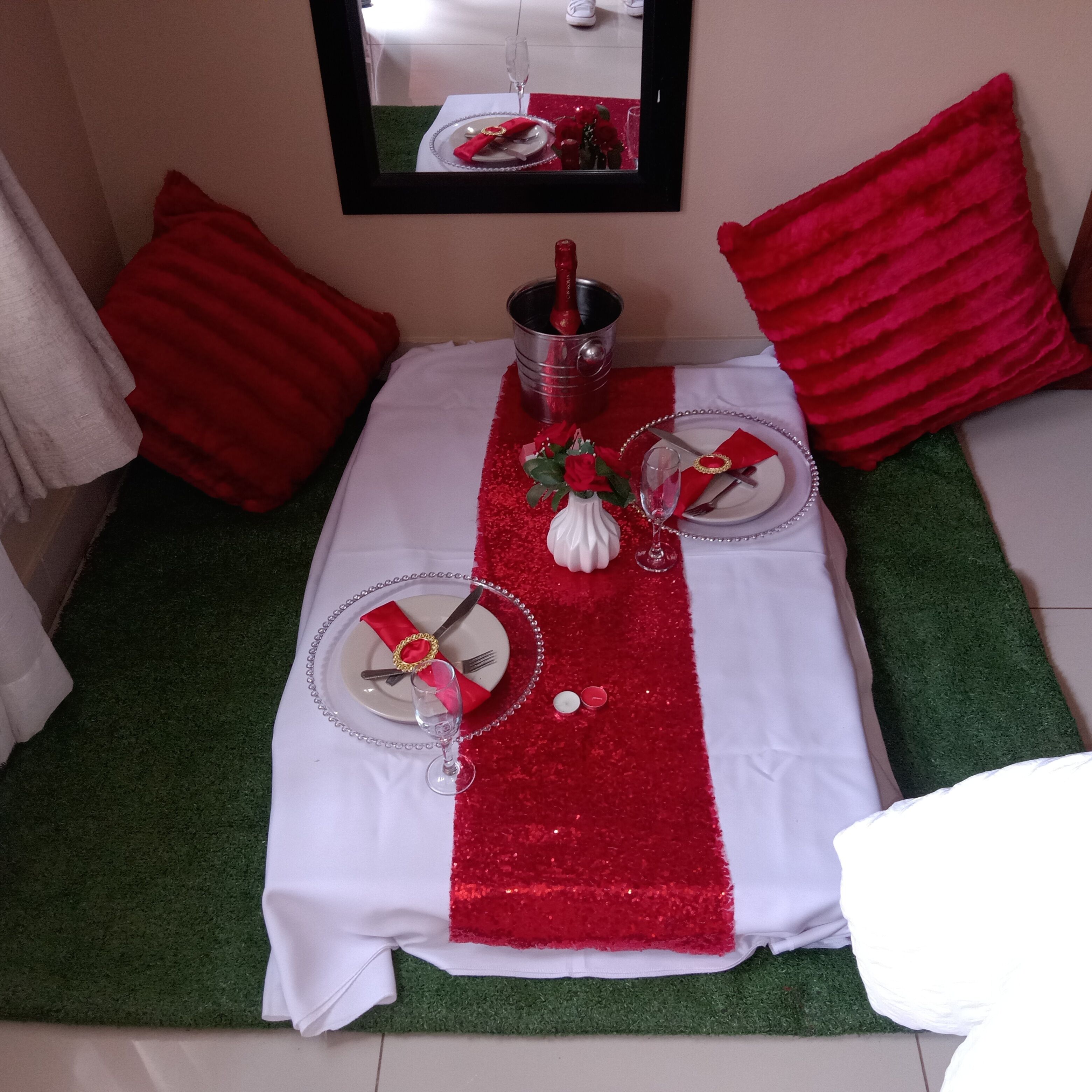 Una mesa de comedor bien dispuesta con camino de mesa rojo y blanco, dos platos, cubiertos y una botella de champán.