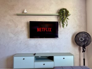 A flat-screen TV mounted on a wall with a shelf above displaying a sailboat model and a potted plant. A black fan is positioned to the right of the TV.