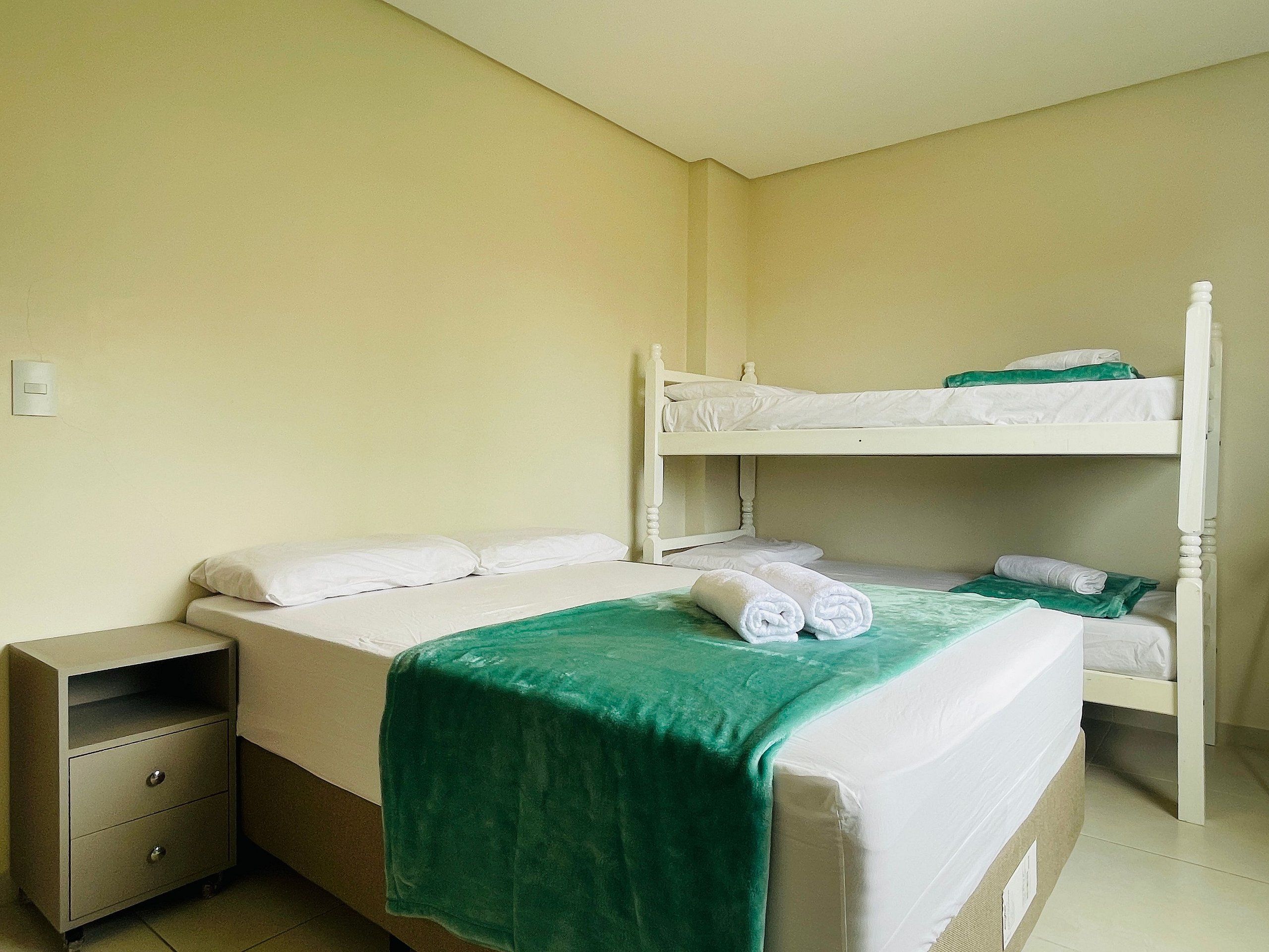 A single bed with a green blanket, a nightstand with drawers, and a bunk bed with white bedding.