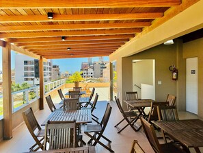 A covered outdoor patio with wooden tables and chairs, a potted plant, and a view of surrounding buildings.