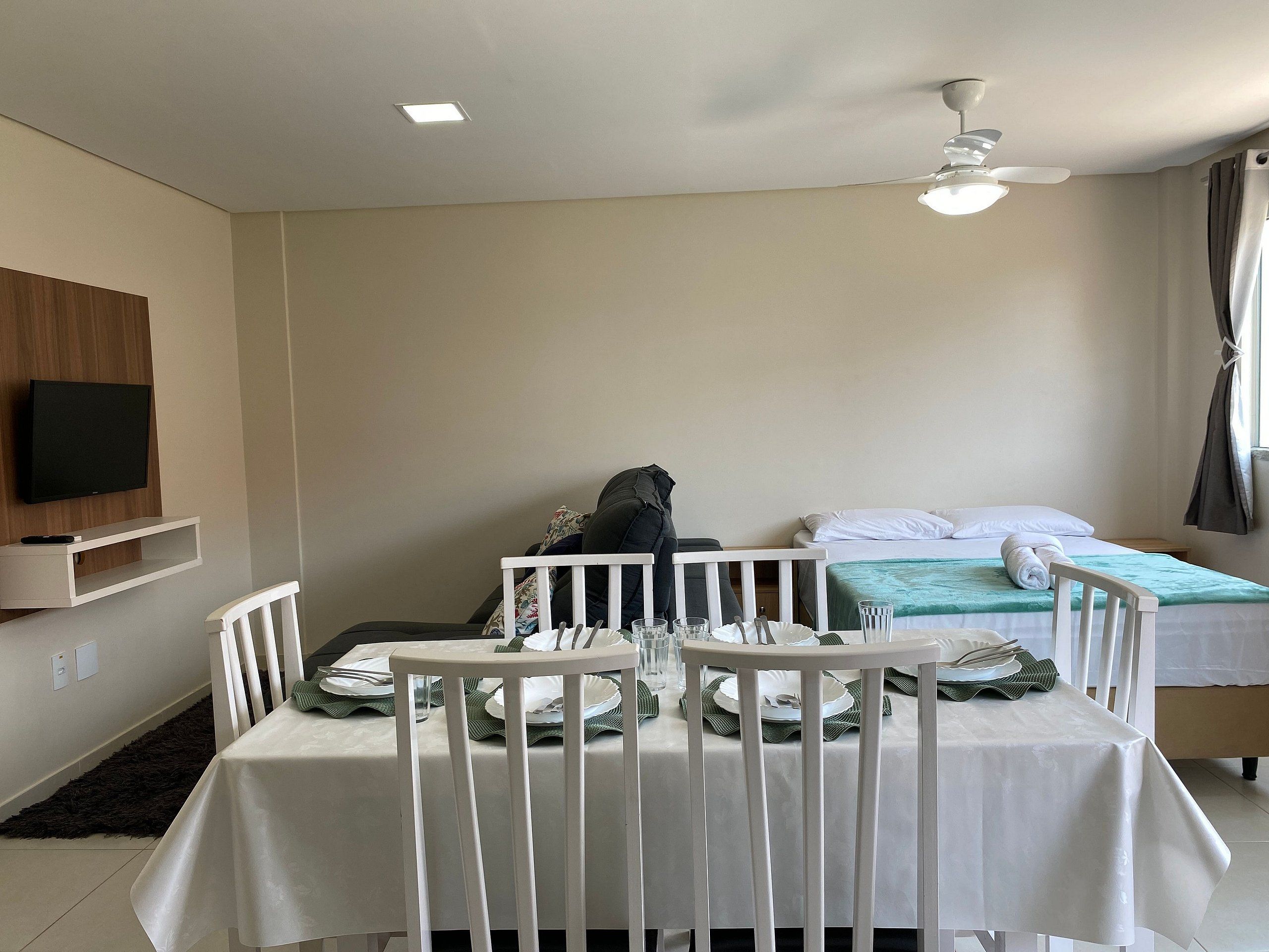 A hotel room with a bed, a dining table set for two, a TV mounted on the wall, and a ceiling fan.