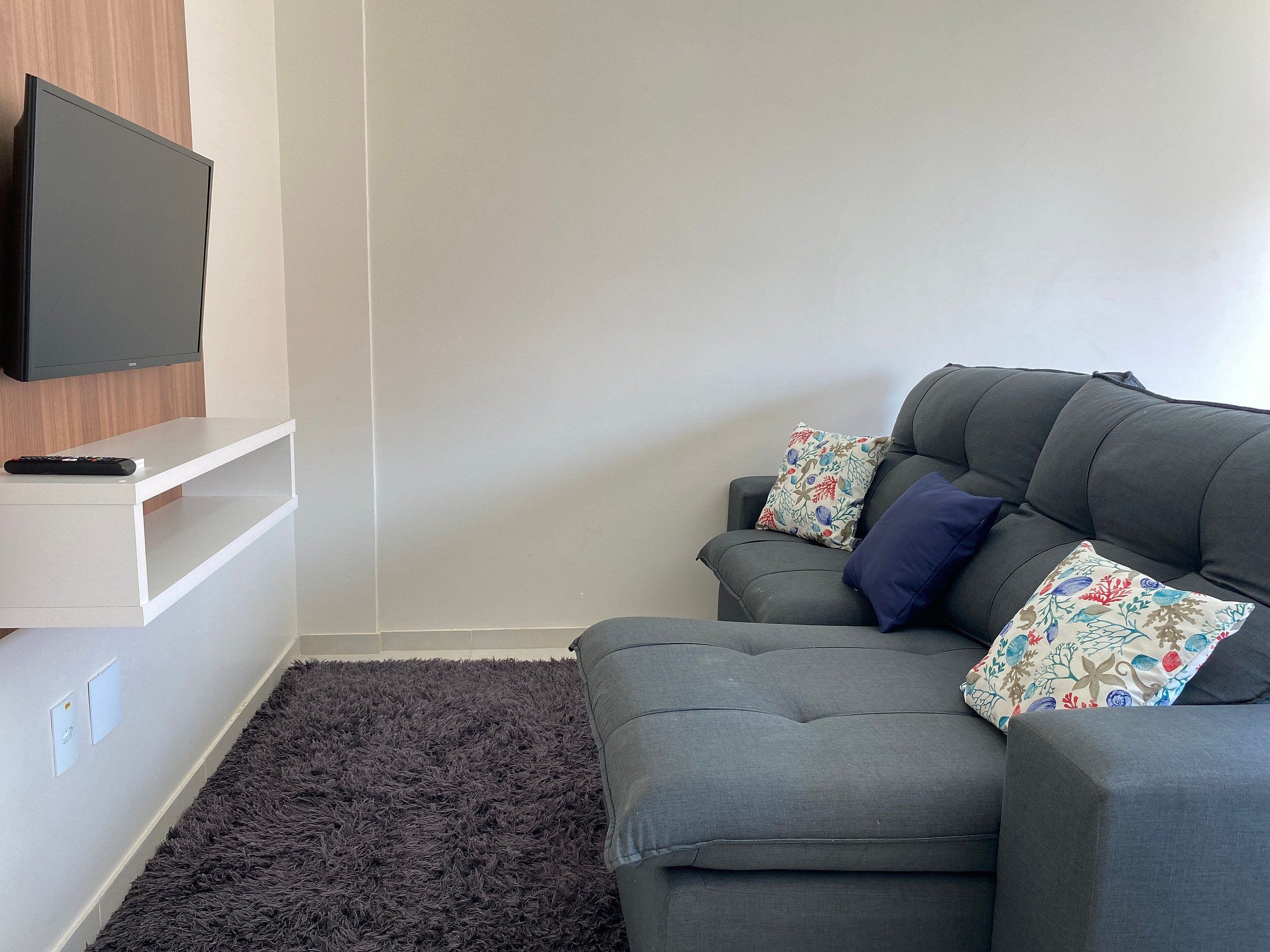 A modern living room with a grey sofa, two patterned cushions, a remote control, and a mounted TV on a white shelf.