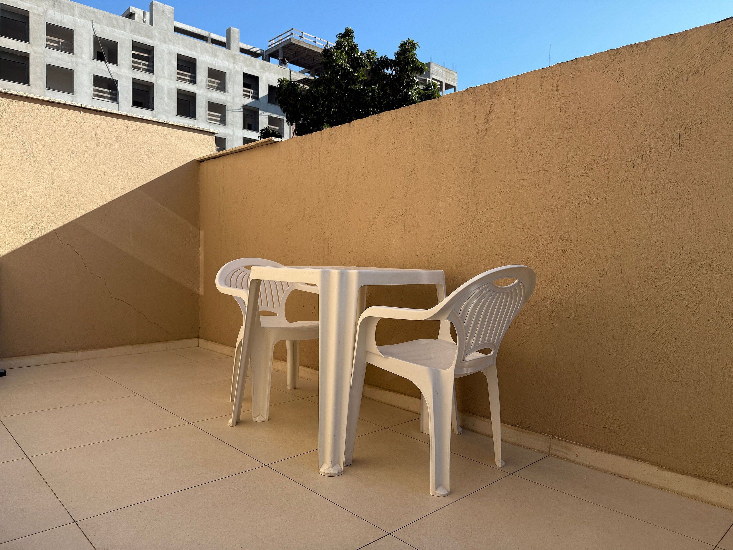 A small outdoor patio area with a white table and two white plastic chairs, a tiled floor, and a beige wall.