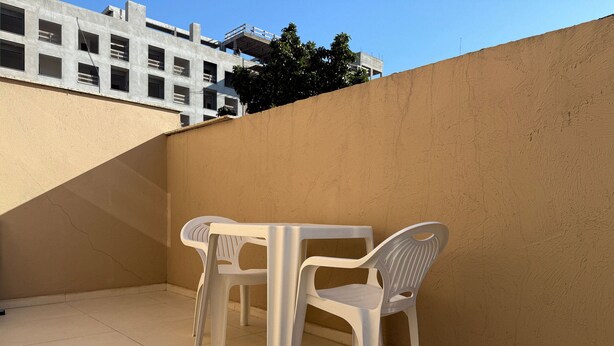 A small outdoor patio area with a white table and two white plastic chairs, a tiled floor, and a beige wall. - Maria Julia (Bombinhas)