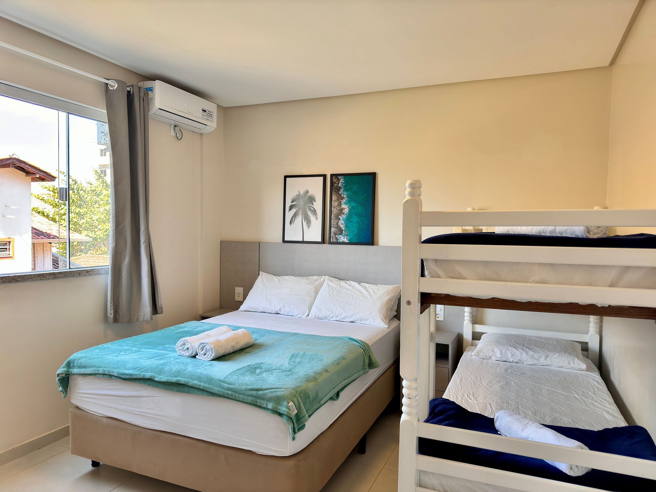 A hotel room with a bed, a bunk bed, a window with curtains, an air conditioning unit, and a framed picture on the wall.