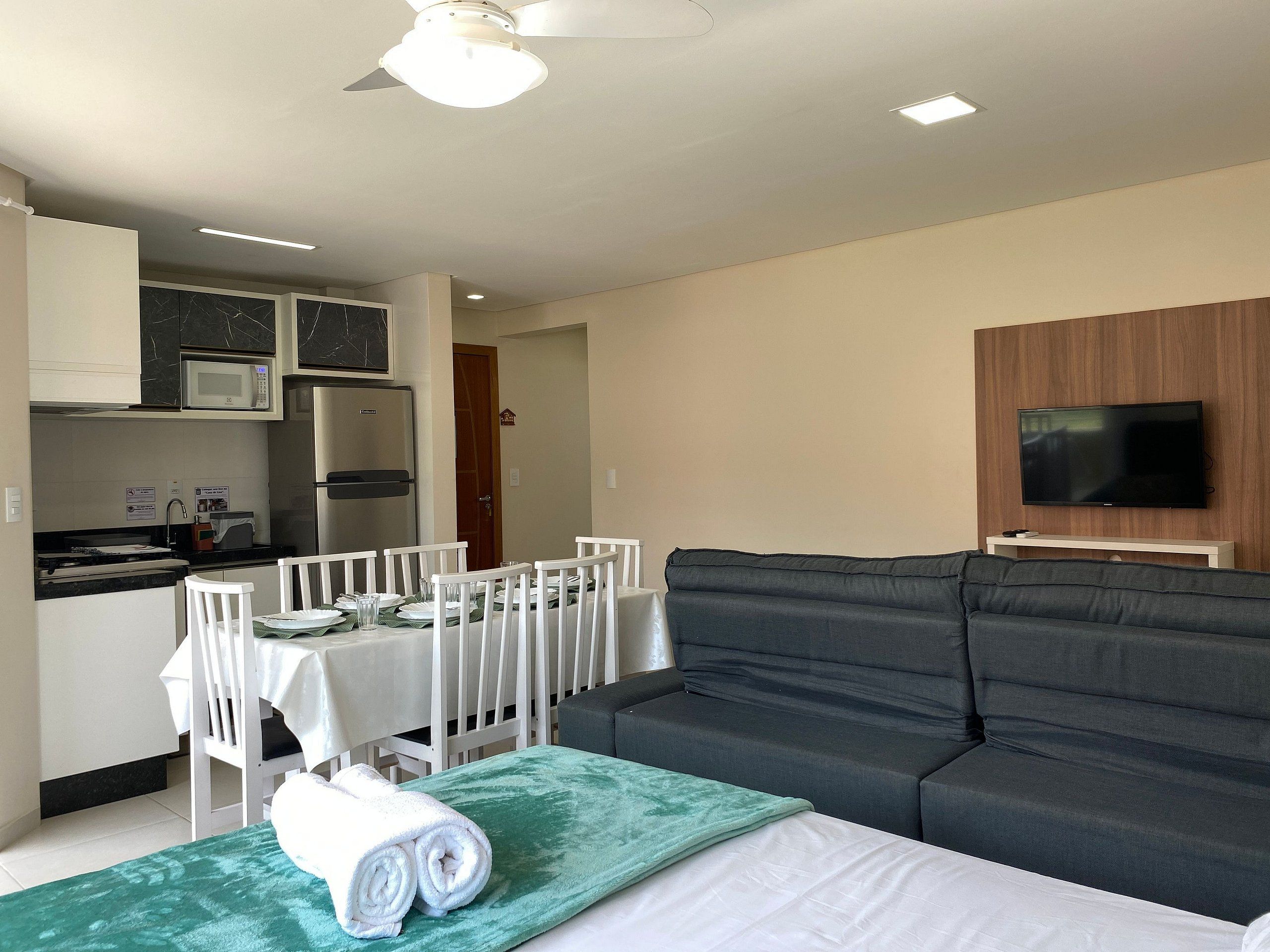 Un salón moderno con un sofá gris, una mesa de comedor con sillas blancas, un televisor de pantalla plana montado en un panel de madera y un ventilador de techo.