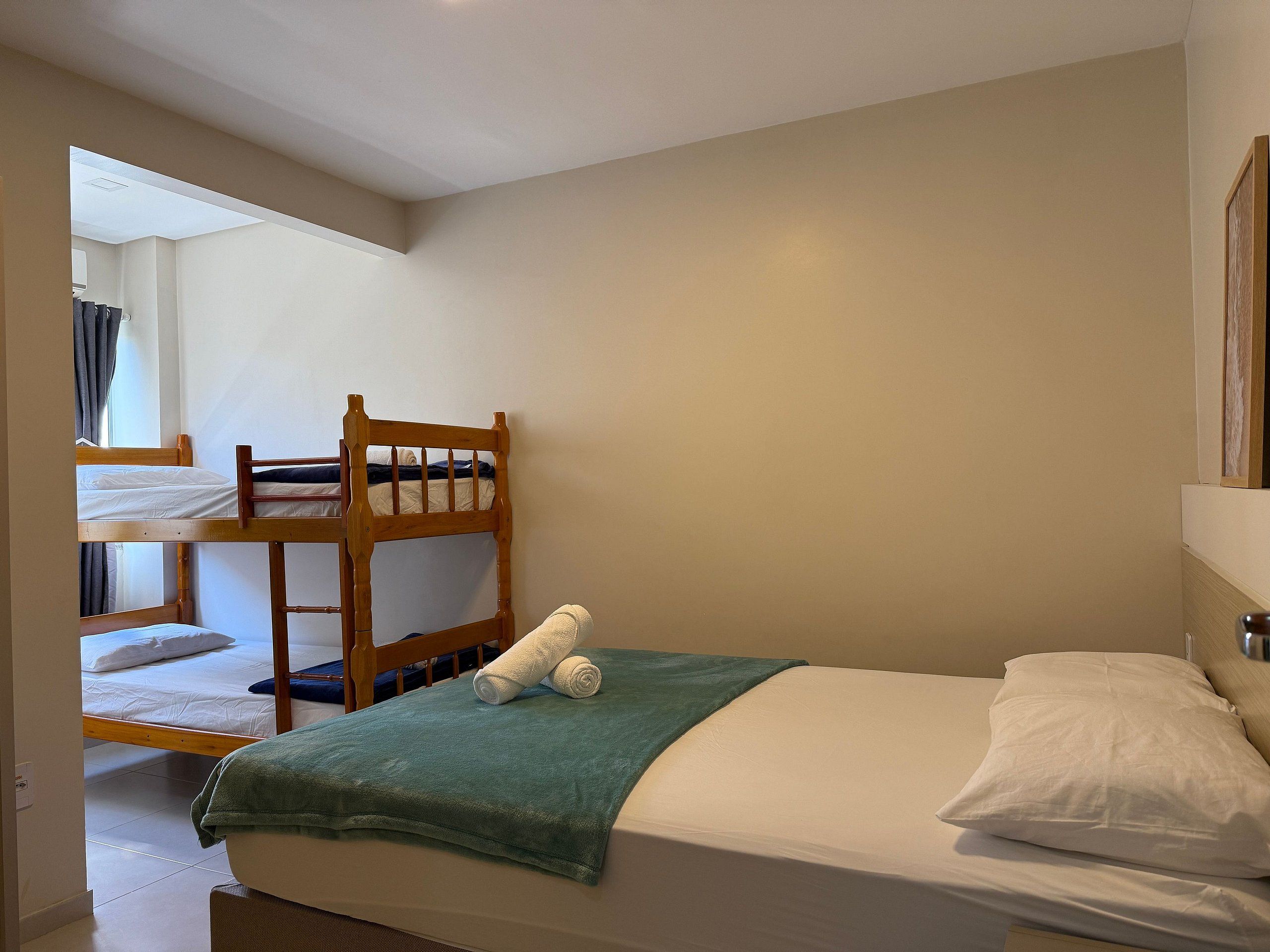 Un lit simple avec une couverture verte et des oreillers blancs, un lit superposé en bois à l’arrière-plan et un miroir au mur.
