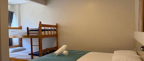 Un lit simple avec une couverture verte et des oreillers blancs, un lit superposé en bois à l’arrière-plan et un miroir au mur.