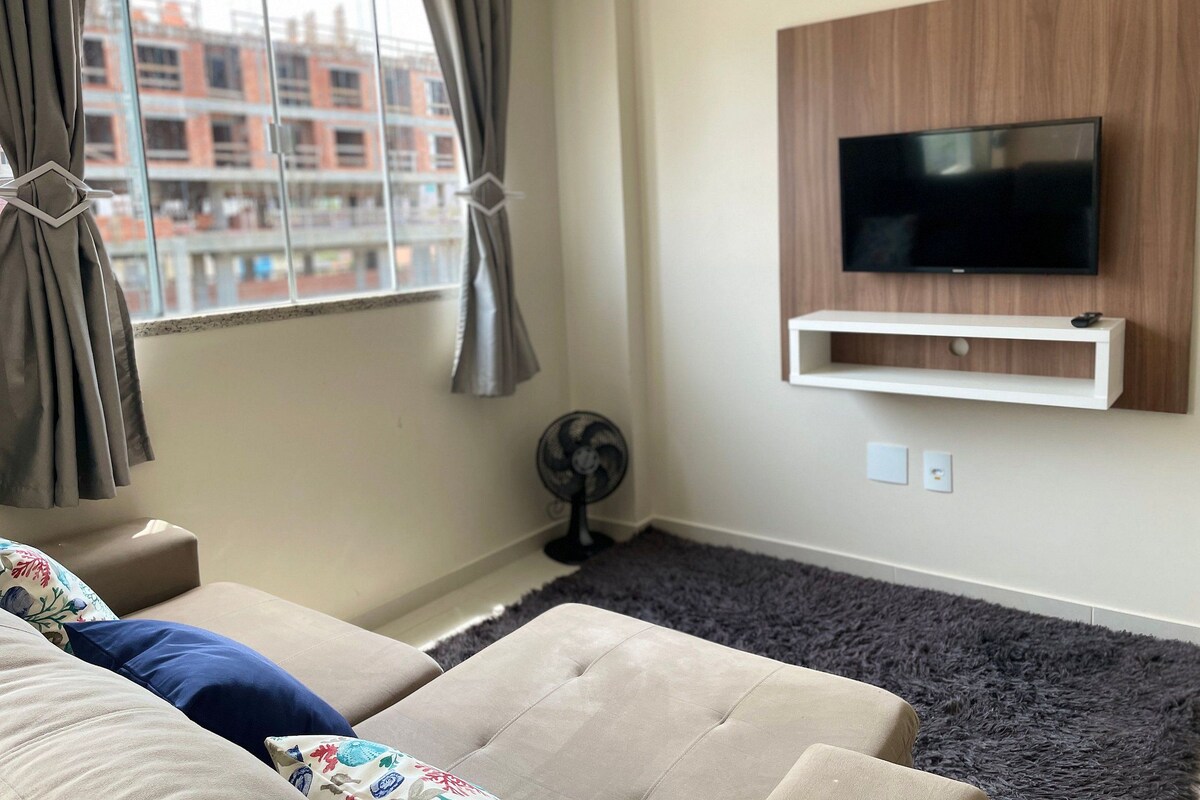 A living room with a sofa, a TV mounted on the wall, a window with curtains, and a fan.