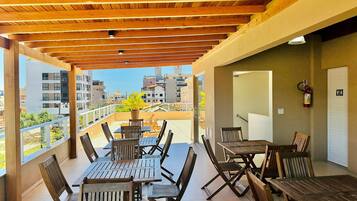 A covered outdoor patio with wooden tables and chairs, a potted plant, and a view of surrounding buildings.