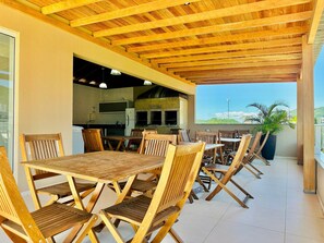 An outdoor dining area with wooden tables and chairs, a kitchen in the background, and a view of the landscape. - Maria Julia (Bombinhas)