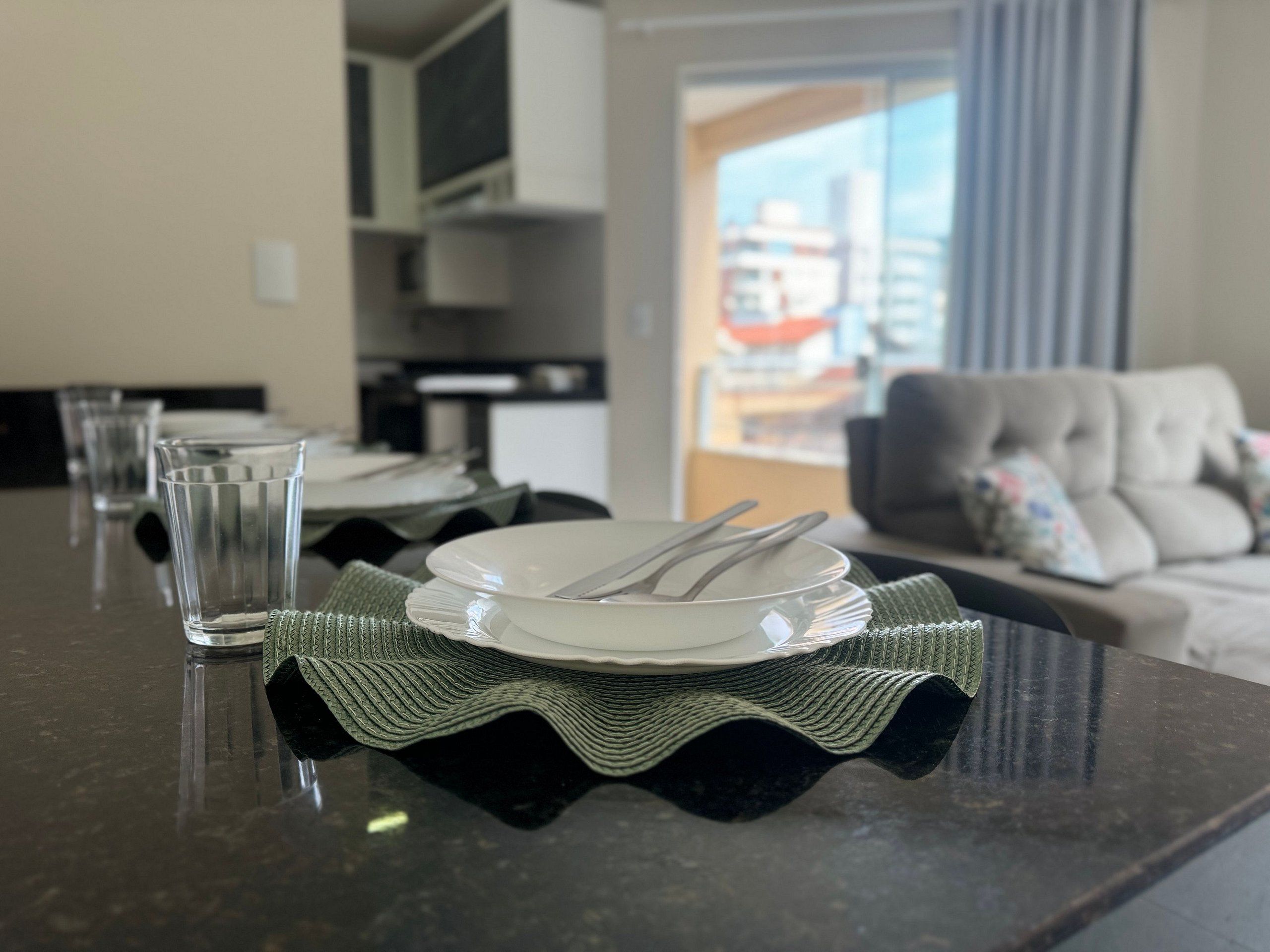 A dining table set with plates, cutlery, and glasses, with a view of a living room and a cityscape through a window.