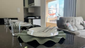 A dining table set with plates, cutlery, and glasses, with a view of a living room and a cityscape through a window.