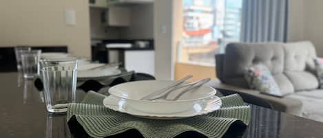 A dining table set with plates, cutlery, and glasses, with a view of a living room and a cityscape through a window.