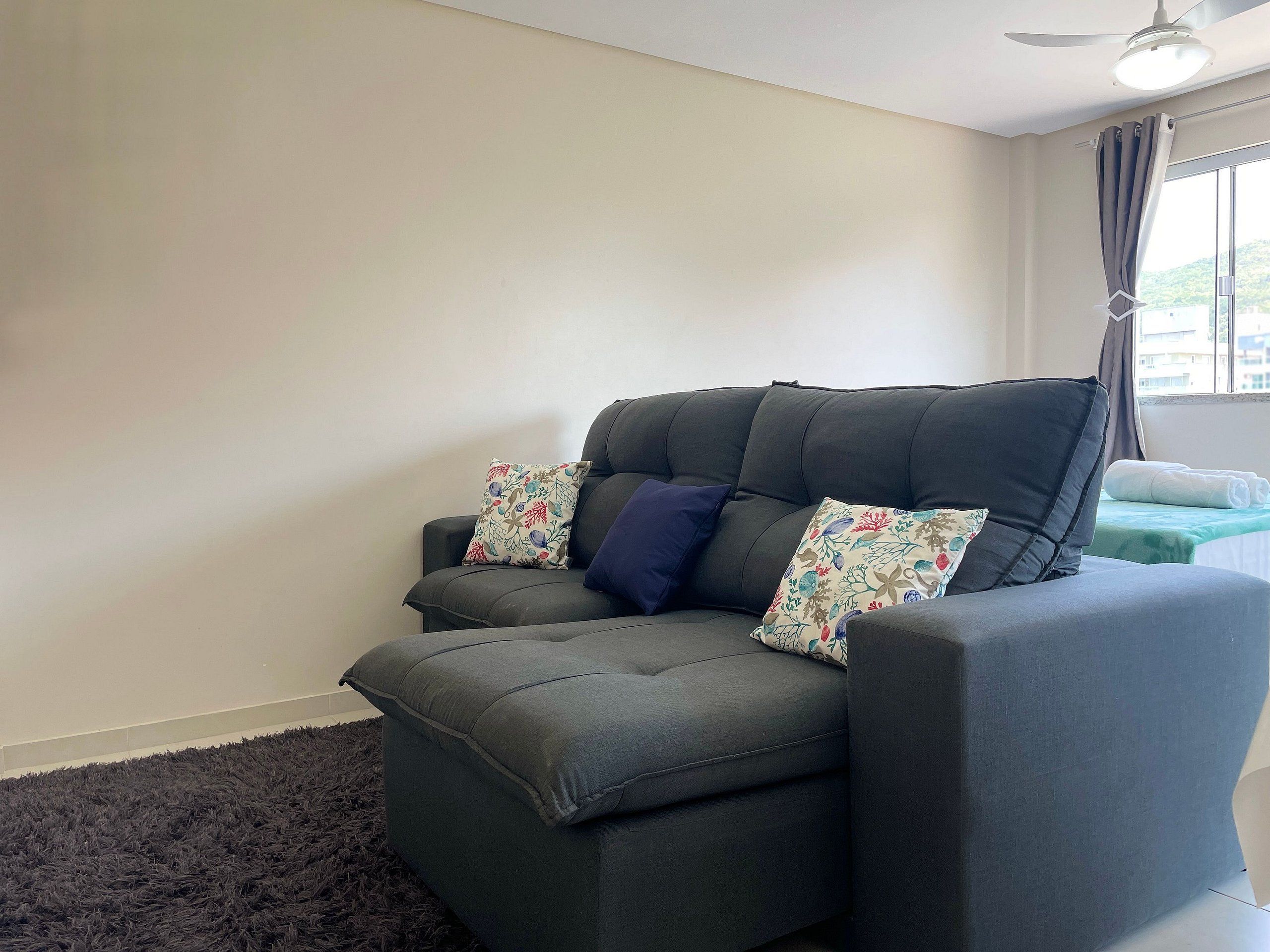A grey sofa with floral cushions, a dark rug, and a ceiling fan in a room with plain walls and a window with curtains.