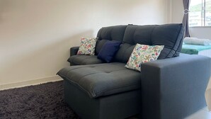 A grey sofa with floral cushions, a dark rug, and a ceiling fan in a room with plain walls and a window with curtains. - Maria Julia (Bombinhas)