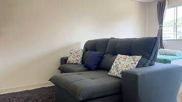 A grey sofa with floral cushions, a dark rug, and a ceiling fan in a room with plain walls and a window with curtains.