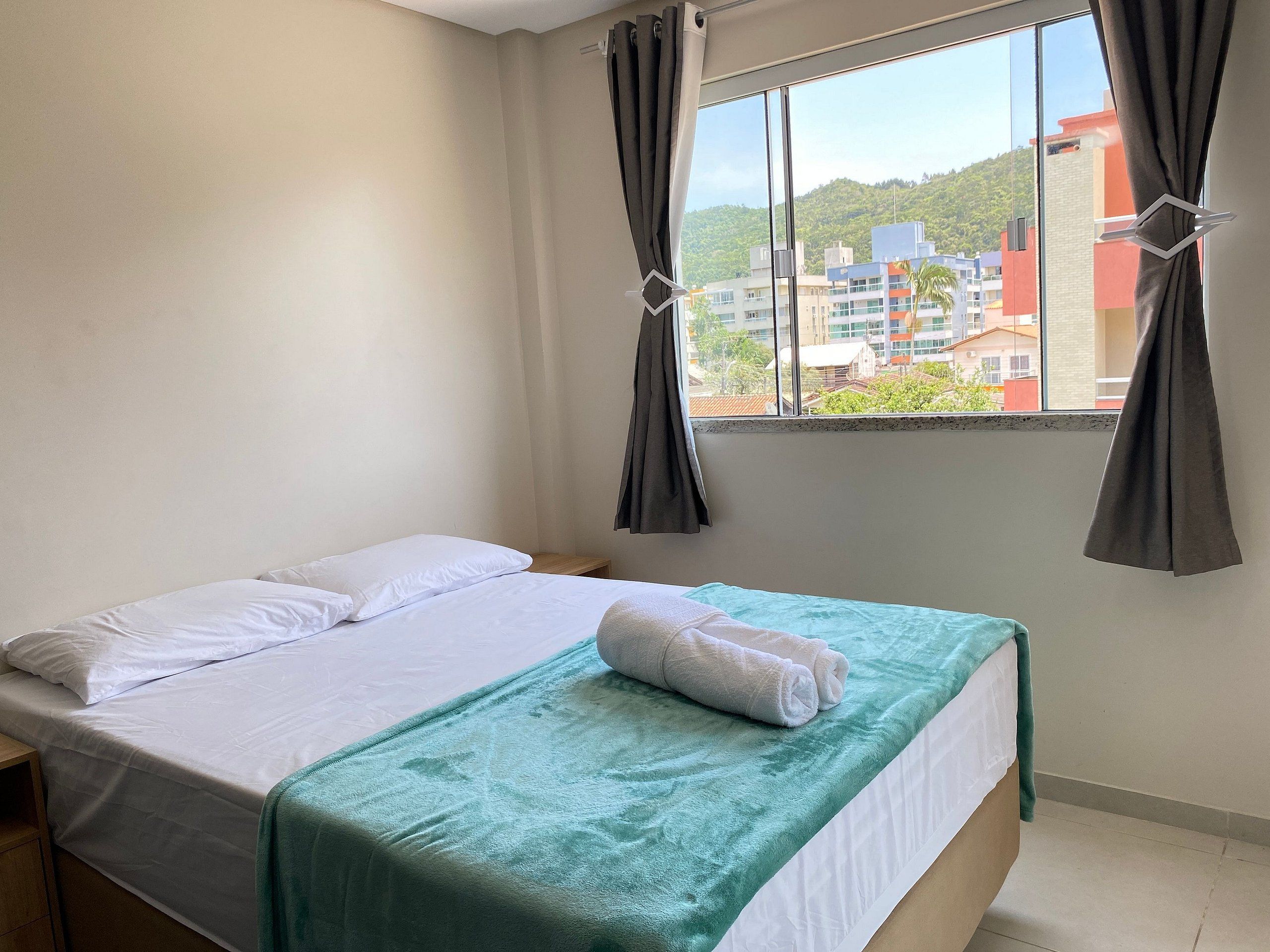 A single bed with white linens, a teal blanket, and a folded towel. A window with curtains is visible, offering a view of buildings and greenery.